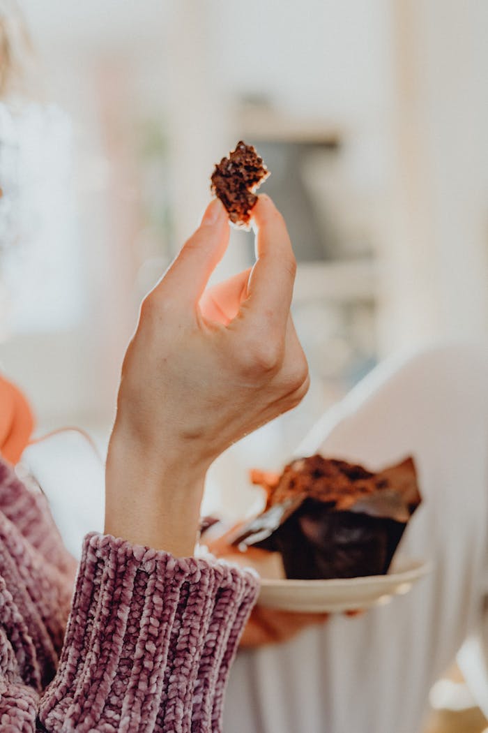 A close-up view of a hand holding a piece of chocolate cake with a cozy background.